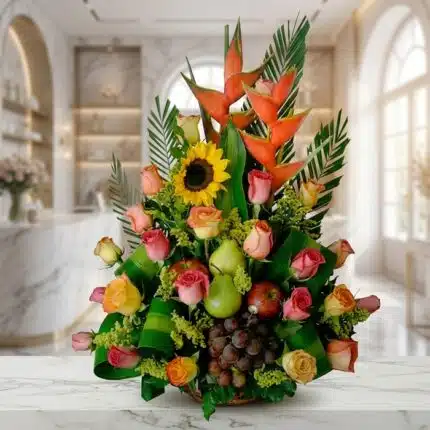 Arreglo floral con rosas de colores, girasoles, heliconias, uvas, peras y manzanas, sobre una superficie de mármol en un ento