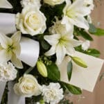 Primer plano macro de un arreglo floral fúnebre blanco con lirios, rosas y claveles, gotas de rocío, cinta satinada y un sobr