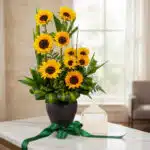 Arreglo floral GIULIANA con girasoles radiantes en jarrón oscuro, sobre mármol, con tarjeta de regalo y cinta verde satinada,