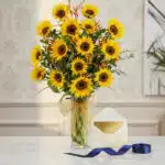 Arreglo floral de girasoles LENU en jarrón de cristal con lazo dorado, tarjeta de regalo elegante y cinta azul sobre mesa de