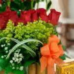 Primer plano extremo de Arreglo Floral Musca con rosas rojas, follaje verde, gotas de rocío y lazo naranja, destacando textur