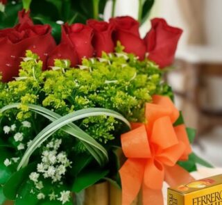 Primer plano extremo de Arreglo Floral Musca con rosas rojas, follaje verde, gotas de rocío y lazo naranja, destacando textur