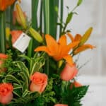 Primer plano extremo de un arreglo floral 'Suspiro' con lirios naranjas vibrantes, rosas color salmón y follaje verde, destac