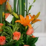 Primer plano extremo de un arreglo floral 'Suspiro' con lirios naranjas vibrantes, rosas color salmón y follaje verde, destac