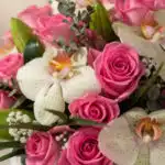 Primer plano extremo de un arreglo floral con rosas rosadas, orquídeas blancas con gotas de rocío, y follaje verde, destacand