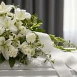 Primer plano de un arreglo fúnebre cubre caja Asunción con lirios blancos, claveles y follaje verde, con rocío sutil y cinta