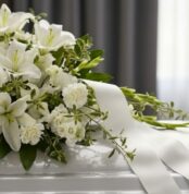 Primer plano de un arreglo fúnebre cubre caja Asunción con lirios blancos, claveles y follaje verde, con rocío sutil y cinta