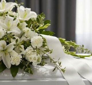 Primer plano de un arreglo fúnebre cubre caja Asunción con lirios blancos, claveles y follaje verde, con rocío sutil y cinta