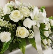 Arreglo fúnebre cubre caja Bondad con rosas blancas, lirios y claveles, follaje verde y cinta satinada, en una fotografía mac