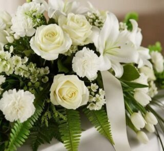Arreglo fúnebre cubre caja Bondad con rosas blancas, lirios y claveles, follaje verde y cinta satinada, en una fotografía mac