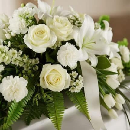 Arreglo fúnebre cubre caja Bondad con rosas blancas, lirios y claveles, follaje verde y cinta satinada, en una fotografía mac