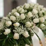 Fotografía macro premium del Arreglo Fúnebre Cubre Caja Cielo, con rosas blancas, follaje verde y rocío sutil. Detalle de cin