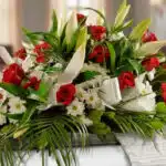 Arreglo fúnebre cubre caja Paraíso con rosas rojas, lirios blancos y margaritas, con follaje verde y cinta satinada.