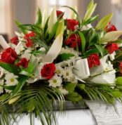 Arreglo fúnebre cubre caja Paraíso con rosas rojas, lirios blancos y margaritas, con follaje verde y cinta satinada.