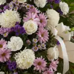 Fotografía macro premium de arreglo fúnebre con rosas blancas, crisantemos y margaritas lilas, rocío sutil, cinta satinada y