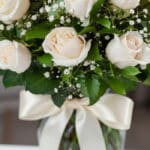 Fotografía macro premium de un arreglo fúnebre con rosas blancas, follaje verde con rocío sutil y un lazo de cinta satinada c