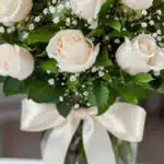 Fotografía macro premium de un arreglo fúnebre con rosas blancas, follaje verde con rocío sutil y un lazo de cinta satinada c