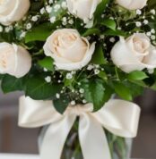 Fotografía macro premium de un arreglo fúnebre con rosas blancas, follaje verde con rocío sutil y un lazo de cinta satinada c