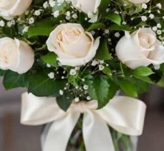 Fotografía macro premium de un arreglo fúnebre con rosas blancas, follaje verde con rocío sutil y un lazo de cinta satinada c