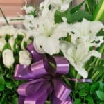 Primer plano extremo de un arreglo fúnebre Pureza con lirios blancos, rosas blancas y un lazo morado, mostrando texturas hipe