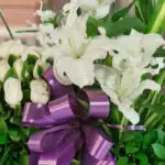 Primer plano extremo de un arreglo fúnebre Pureza con lirios blancos, rosas blancas y un lazo morado, mostrando texturas hipe