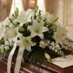 Arreglo fúnebre de rosas blancas y lirios con rocío sutil, cinta satinada y borde de sobre de condolencias. Texturas hiperrea