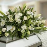 Fotografía macro de un arreglo fúnebre cubre caja con rosas blancas, lirios y follaje verde, con rocío sutil y cinta satinada