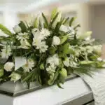 Fotografía macro de un arreglo fúnebre cubre caja con rosas blancas, lirios y follaje verde, con rocío sutil y cinta satinada