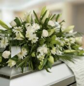 Fotografía macro de un arreglo fúnebre cubre caja con rosas blancas, lirios y follaje verde, con rocío sutil y cinta satinada