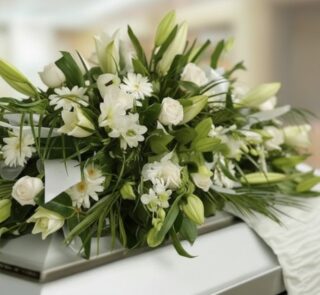 Fotografía macro de un arreglo fúnebre cubre caja con rosas blancas, lirios y follaje verde, con rocío sutil y cinta satinada