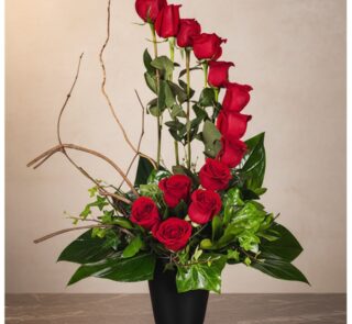 Arreglo floral GIANNA con 13 rosas rojas frescas en un jarrón negro, ramas decorativas y follaje verde, sobre una superficie