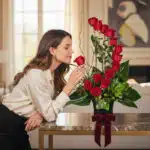 Mujer elegante oliendo delicadamente una rosa roja de un arreglo floral GIANNA con 13 rosas, en un lujoso interior de hogar.