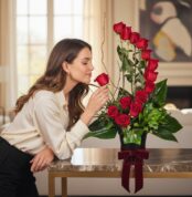 Mujer elegante oliendo delicadamente una rosa roja de un arreglo floral GIANNA con 13 rosas, en un lujoso interior de hogar.