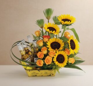 Arreglo floral vibrante con girasoles, rosas naranjas y cuatro botellas de cerveza Corona en una base amarilla, ideal para ce