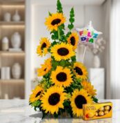 Arreglo floral exótico con girasoles grandes y vibrantes, follaje verde, un globo de 'Feliz Día' y una caja de chocolates Fer