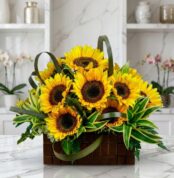 Arreglo floral exótico con girasoles Soleil grandes y abiertos, hojas verdes vibrantes, en una cesta de madera oscura sobre m