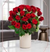 Arreglo floral Jianna con 25 rosas rojas frescas en un elegante jarrón blanco sobre mesa de mármol, fondo difuminado de sala