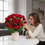 Mujer elegante admirando con deleite un Arreglo Jianna de 25 rosas rojas en un jarrón beige sobre una mesa de mármol, en un h