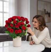 Mujer elegante admirando con deleite un Arreglo Jianna de 25 rosas rojas en un jarrón beige sobre una mesa de mármol, en un h