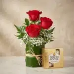 Elegante arreglo floral con tres rosas rojas vibrantes, follaje verde y flores blancas, acompañado de una caja de bombones Fe