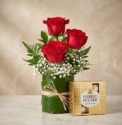 Elegante arreglo floral con tres rosas rojas vibrantes, follaje verde y flores blancas, acompañado de una caja de bombones Fe