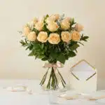 Bouquet de 12 rosas Celestina color melocotón, atado con cinta de satén, junto a una tarjeta de regalo elegante en un sobre a