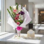 Bouquet de flores GIA con gerberas rosadas, lirios blancos y flores moradas, envuelto en papel blanco y rosa, con cinta fucsi