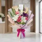 Bouquet de Flores GIA con gerberas rosadas, lirios blancos y flores moradas, envuelto en papel coreano elegante y lazo fucsia