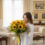 Mujer joven y elegante con camisa blanca admira un gran bouquet de girasoles frescos y radiantes, atado con un lazo de yute,