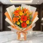 Bouquet LIRICA con lirios naranjas vibrantes, rosas amarillas abiertas y gerberas rojas, envuelto en papel coreano elegante s