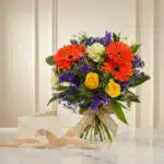 Bouquet Floral SABRINA con rosas amarillas, gerberas naranjas, flores moradas y follaje verde, atado con cinta de yute, sobre