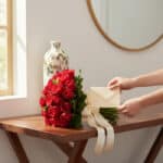 Manos delicadas colocando una tarjeta en un sobre junto a un exuberante bouquet TIZIANA de 80 rosas rojas premium, atado con