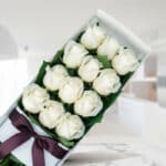 Caja de 12 rosas blancas frescas y abiertas con un moño elegante, sobre una superficie de mármol en un entorno de floristería