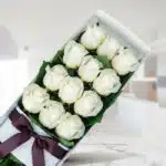 Caja de 12 rosas blancas frescas y abiertas con un moño elegante, sobre una superficie de mármol en un entorno de floristería